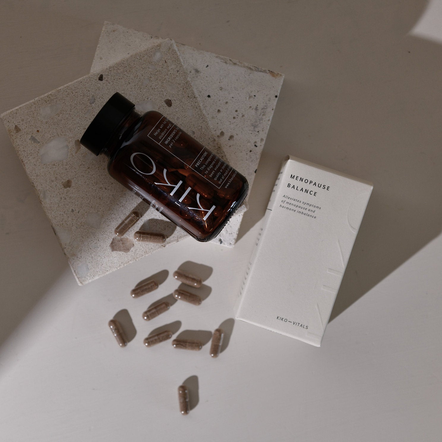 A bottle labeled 'MENOPAUSE BALANCE' with several tablets spilling out in front of it, alongside a white box with the product name and a few tablets visible on it. The items are placed on a light surface with a shadow falling over part of the image.