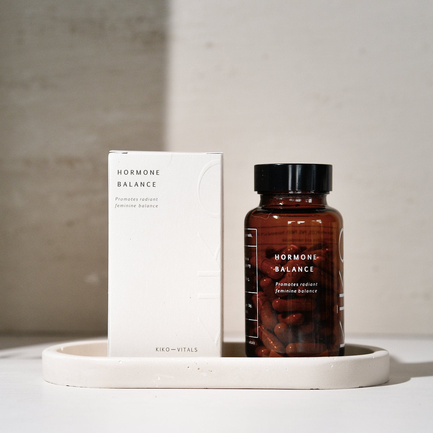 A product named 'Hormone Balance' by Kiko Vitals, consisting of a glass bottle with capsules inside and a white box with text. The bottle and box are placed on a white surface with a shadow falling over the background.