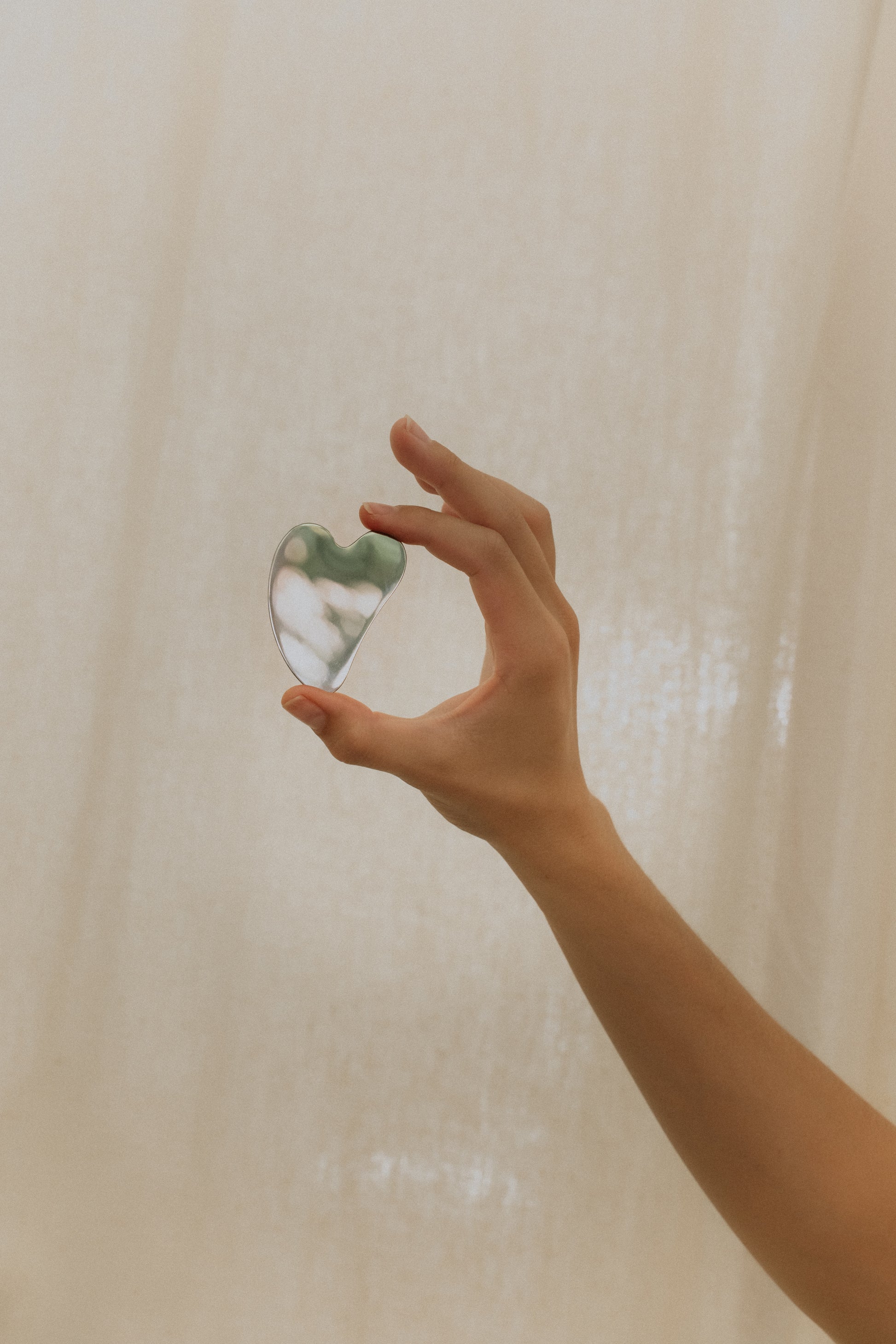 A person's hand holding a small chrome Gua Sha tool against a neutral background.