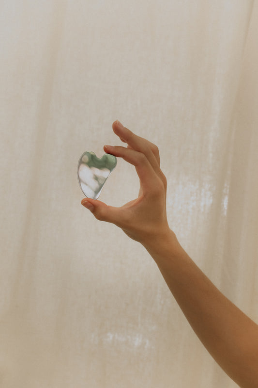 A person's hand holding a small chrome Gua Sha tool against a neutral background.