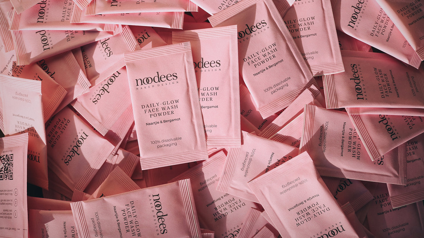 A pile of dissolvable pink sachets labeled 'noodees daily glow face polish powder', designed for facial cleansing.