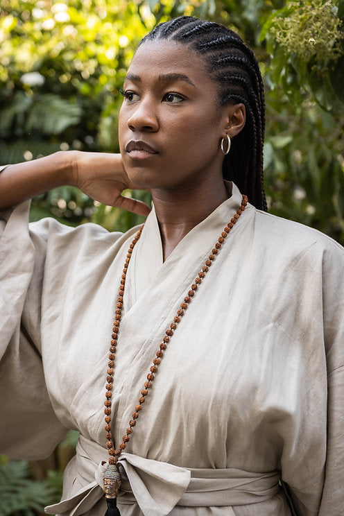 A woman wearing a beige linen cotton blend kimono with full-length kimono-style sleeves and a wide fabric belt. She is also wearing a long beaded necklace.