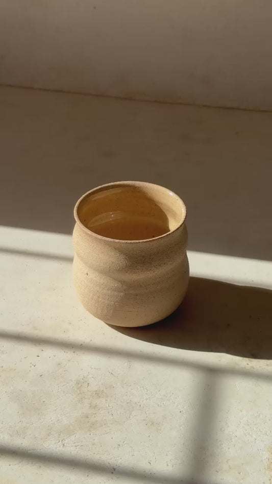 A ceramic mug in a beige color, displayed among other pottery pieces on a table.