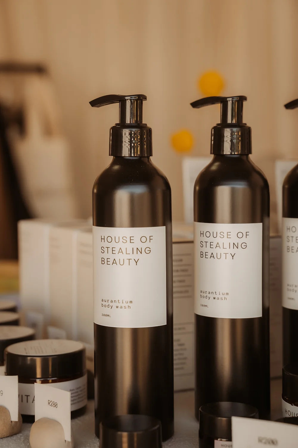 Two black pump bottles labeled 'HOUSE OF STEALING BEAUTY' and 'Aurantium Body Wash', displayed on a table with other skincare products.