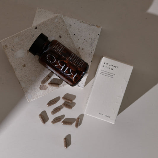 A bottle labeled 'MENOPAUSE BALANCE' with several tablets spilling out in front of it, alongside a white box with the product name and a few tablets visible on it. The items are placed on a light surface with a shadow falling over part of the image.